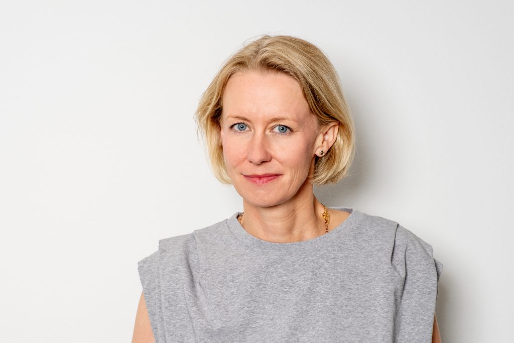 Stefanie Eikel steht vor einer weißen Wand und schaut lächelnd in die Kamera.